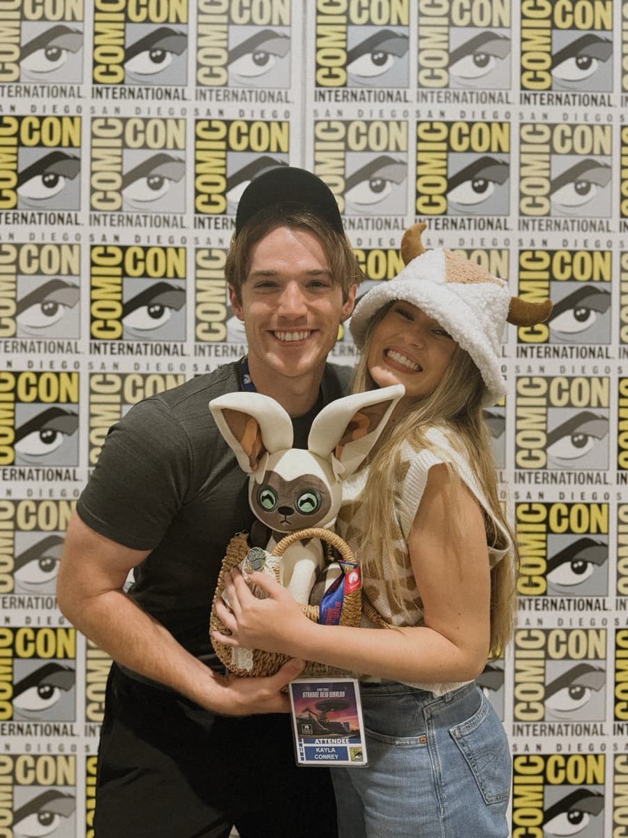 Kayla and her husband holding a Momo plush in front of the Comic-Con backdrop